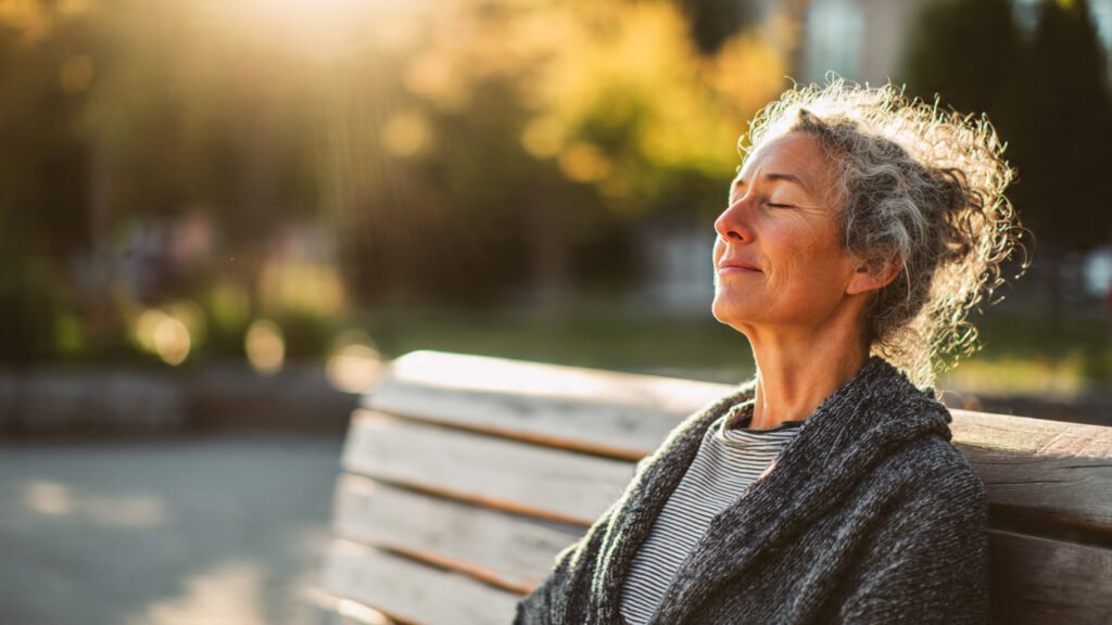 American woman over 45 sitting alone in peaceful park on a bench during early morning, eyes closed practicing deep breathing, casual comfortable clothing, natural sunlight, relaxed environment, realistic photography, 16:9 ratio, no text